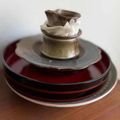 Stacked ceramic and lacquerware dishware, including a small stoneware creamer, saucer, and red-and-black lacquer trays.