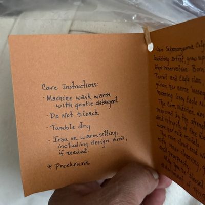 Sale Photo Thumbnail #127: Handwritten care instructions and artist information tag attached to a textile item.