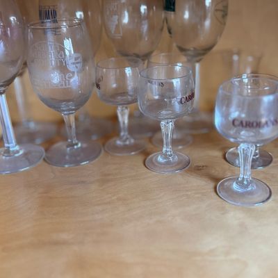 Collection of assorted promotional glassware and cordials on a wooden shelf, including wine glasses and Carolans liqueur glasses.