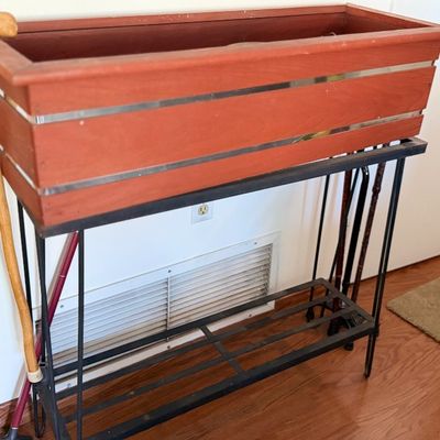 Rectangular wooden planter box with metallic trim set on a black metal wrought-iron stand with a lower slat shelf.