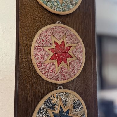 Trio of vintage folded fabric star ornaments framed in wooden embroidery hoops, mounted on a dark wood panel.