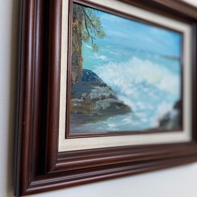 Sale Photo Thumbnail #150: Framed original oil painting depicting a coastal seascape with crashing waves, mounted in a dark wooden frame with a cream canvas liner.