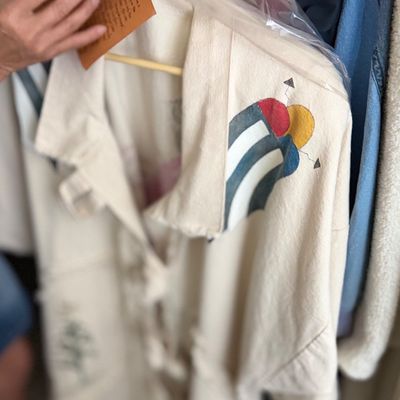 Sale Photo Thumbnail #128: Cream-colored garment featuring colorful graphic art on the shoulder, stored in a clear garment bag with attached tag.