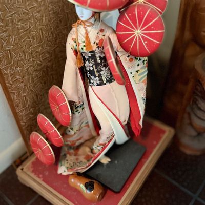 Vintage Japanese geisha doll dressed in a patterned kimono and holding red parasols, mounted on a wooden display base.