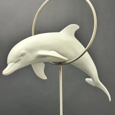 Sale Photo Thumbnail #203: Matte white porcelain dolphin figurine leaping through a metal ring, mounted on a metal stand.