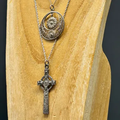 Sale Photo Thumbnail #178: Two silver pendant necklaces displayed on a wooden bust, featuring an ornate Celtic cross and an intricate filigree circular pendant.