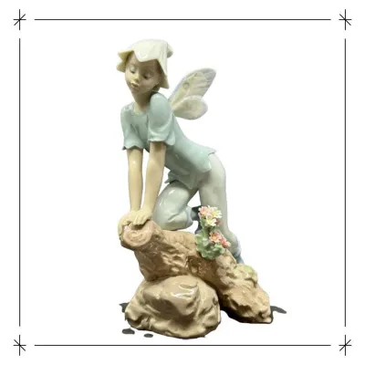 Sale Photo Thumbnail #201: Lladró porcelain fairy figurine depicting a winged sprite perched on a log with delicate flowers, rendered in a muted pastel palette with a glossy finish.