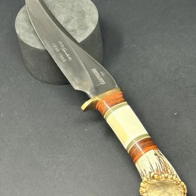 Sale Photo Thumbnail #15: Randall Made 50th Anniversary (1938-1988) commemorative hunting knife featuring a stag and composite handle, brass guard, and etched blade marked number 068.
