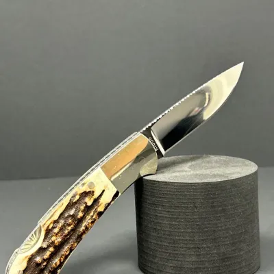 Sale Photo Thumbnail #63: Custom folding pocket knife featuring a polished stainless steel blade, stag horn handle scales, and detailed filework along the spine.