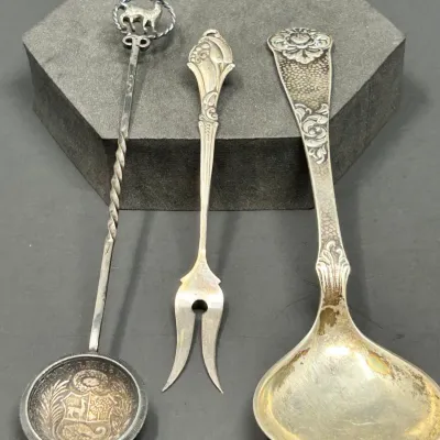 Sale Photo Thumbnail #174: Set of three vintage silver serving utensils, including a coin-bowl spoon with a llama motif, a two-prong pickle fork, and a floral embossed ladle showing slight tarnish.