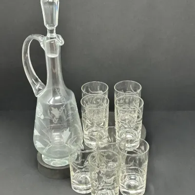 Sale Photo Thumbnail #131: Vintage etched glass decanter with handle and stopper, accompanied by a matching set of eight small drinking glasses featuring floral motifs.