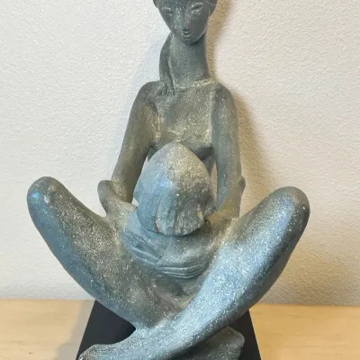 Sale Photo Thumbnail #159: Mid-century modern style sculpture depicting a seated woman holding a child, crafted in a textured gray stone-like material mounted on a black base.