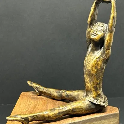 Sale Photo Thumbnail #68: Bronze patina sculpture of a seated young ballerina performing a stretch, mounted on a triangular wooden base.