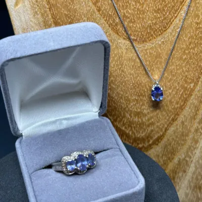 Sale Photo Thumbnail #111: Matching fine jewelry set featuring a three-stone oval tanzanite and diamond ring presented in a velvet box alongside a solitaire tanzanite pendant necklace.