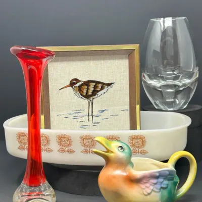 Sale Photo Thumbnail #145: Collection of decorative glassware and ceramics, including a red stretch glass bud vase, a Pyrex Butterfly Gold casserole dish, a framed bird embroidery, and a colorful duck-shaped pitcher.