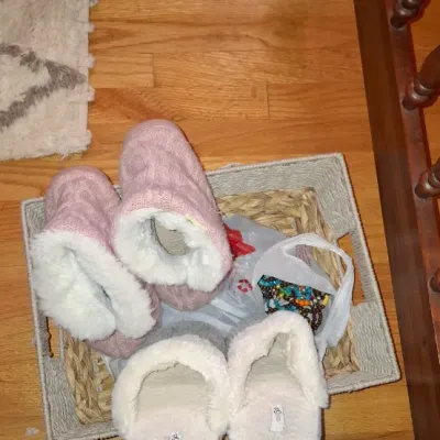 Sale Photo Thumbnail #78: Two pairs of fleece-lined slippers, including pink knit bootie slippers and light grey slip-ons, resting in a shallow woven basket.