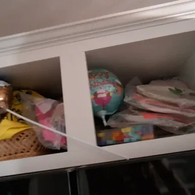 Sale Photo Thumbnail #55: Open white kitchen cabinet filled with party supplies, including helium balloons, paper plates in plastic wrapping, and a woven basket.