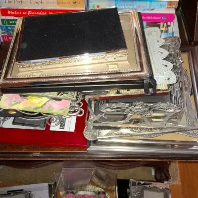 Sale Photo Thumbnail #74: Collection of assorted picture frames including ornate silver-tone metal and floral designs, alongside a stack of paperback fiction books.