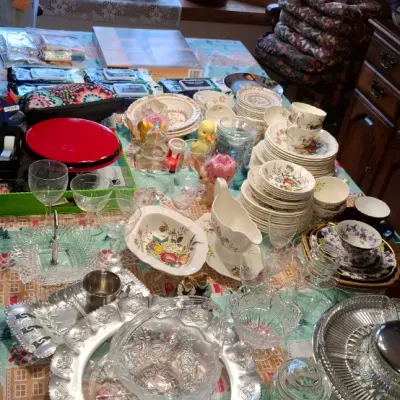 Sale Photo Thumbnail #27: Large assortment of vintage tableware, including floral ceramic dish sets, cut glass serving pieces, silver-tone trays, and stemware, displayed beneath a Tiffany-style stained glass chandelier.