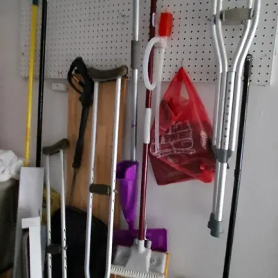 Sale Photo Thumbnail #162: Assorted household items hung on a pegboard, including pairs of aluminum crutches, brooms, and cleaning mops.