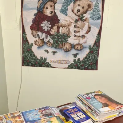 Sale Photo Thumbnail #23: Boyds Bears & Friends woven Christmas wall tapestry hanging on a dark rod above a tabletop display of DVDs and magazines.