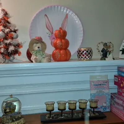 Sale Photo Thumbnail #115: Collection of holiday decor and children's puzzles arranged on a white fireplace mantel, including a small tinsel tree, stacked orange pumpkins, a snow globe, and boxed Barbie puzzles.