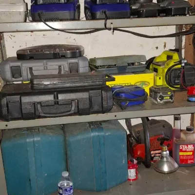 Sale Photo Thumbnail #148: Metal shelving unit filled with various power tools, battery chargers, hard carrying cases, a Sportsman 2-burner propane stove box, and gardening pruners.
