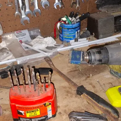 Sale Photo Thumbnail #143: A bright yellow Ryobi cordless drill sitting on a cluttered workbench alongside a red drill bit organizer, hand tools, and a pegboard with wrenches.