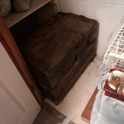 Sale Photo Thumbnail #103: Antique wooden steamer trunk tucked in a closet beneath built-in shelving, showing heavy wear and a distressed dark finish.