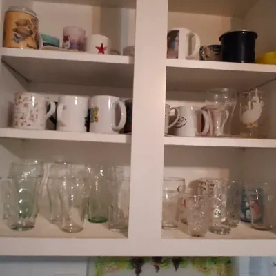 Sale Photo Thumbnail #54: Collection of assorted ceramic mugs and glassware arranged inside a white kitchen cabinet.