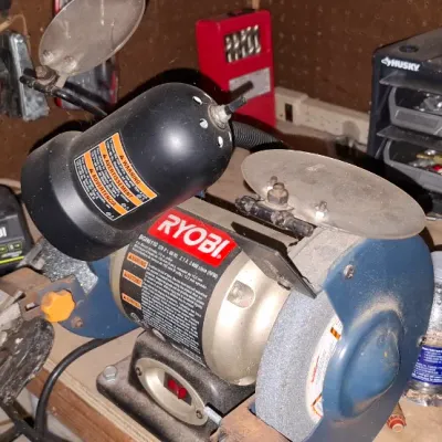 Sale Photo Thumbnail #145: Ryobi 6-inch bench grinder with attached work light and eye shields, alongside a lime green Ryobi cordless drill on a cluttered workbench.