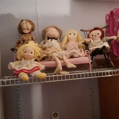 Sale Photo Thumbnail #14: Collection of vintage rag and cloth dolls, including a Raggedy Ann doll and blonde cloth dolls, displayed on a metal wire shelf.