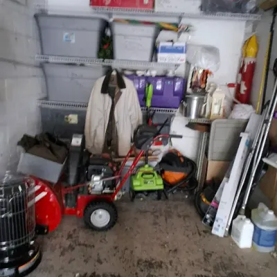 Sale Photo Thumbnail #153: Garage storage area featuring a red snowblower, portable kerosene heater, plastic storage totes, electric lawn equipment, and various household goods.