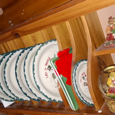 Sale Photo Thumbnail #30: Wooden wall-mounted plate rack filled with holiday-themed 'Let it Snow' snowman dinnerware, accompanied by a snow globe and decorative figurines.