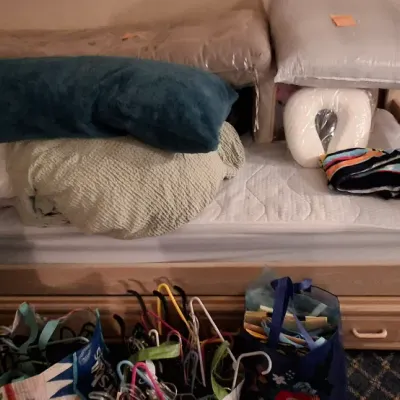 Sale Photo Thumbnail #130: Light wood twin daybed with built-in trundle drawers, stacked with various pillows, linens, and travel pillows, accompanied by shopping bags of clothes hangers on the floor.