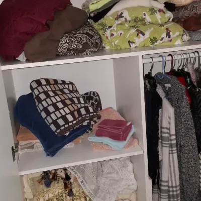 Sale Photo Thumbnail #18: White wooden storage armoire filled with assorted linens, blankets, throw pillows, and hanging clothing, topped with glassware and a novel stag clock.