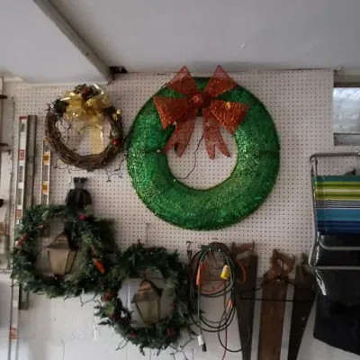 Sale Photo Thumbnail #156: Collection of holiday decorations and garage storage items hanging on pegboard, including artificial Christmas wreaths, a decorative wooden sled, extension cords, hand saws, and a folding lawn chair.