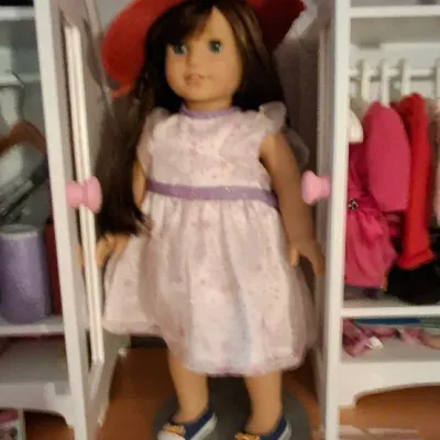 Sale Photo Thumbnail #10: American Girl doll with long brown hair, wearing a red wide-brimmed hat, floral print dress, and blue shoes, displayed in front of doll armoires.
