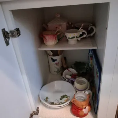 Sale Photo Thumbnail #51: Open white kitchen cabinet filled with assorted decorative tableware, including novelty tea cups, ceramic dishes, a bear-shaped mug, and kitchen tools.