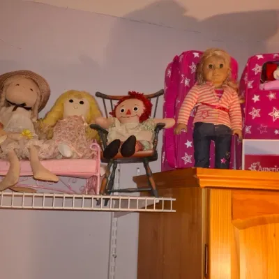 Sale Photo Thumbnail #67: Collection of vintage and modern dolls including an American Girl doll with carrying case, a small Raggedy Ann doll in a mini chair, and various cloth rag dolls displayed on high shelving.