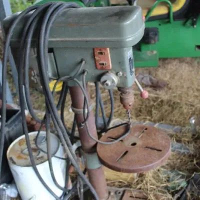 Vintage floor-standing drill press with a green metal casing and rusted circular worktable, showing signs of heavy age and wear.