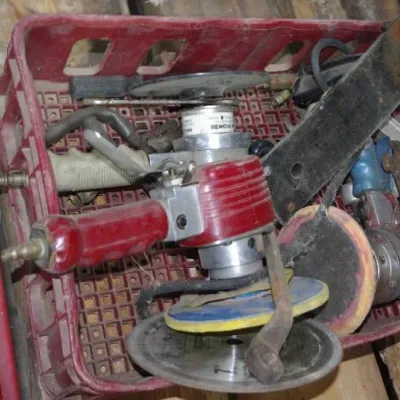 Red plastic crate filled with assorted vintage pneumatic power tools, grinding discs, sanding pads, and metal brackets showing signs of...