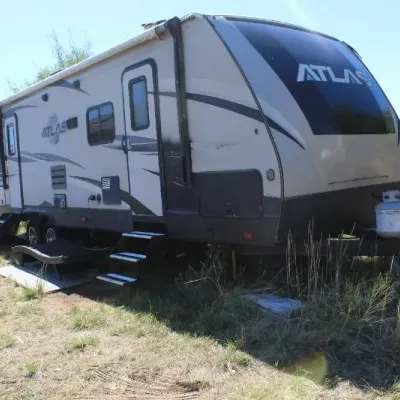Dutchmen Atlas travel trailer camper parked in a grassy field, featuring dual entry steps, fold-out awning, and front propane tanks.