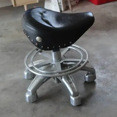 Vintage saddle-style rolling stool featuring a studded black leather seat and a polished aluminum base with an integrated footrest ring.