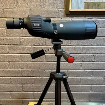 Sale Photo Thumbnail #29: Bushnell spotting scope mounted on an adjustable metal tripod, appearing in good condition.