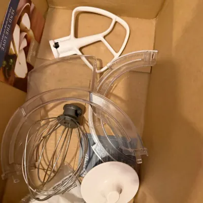 Sale Photo Thumbnail #27: Assorted KitchenAid stand mixer attachments inside a cardboard box, including a wire whip, flat beater, dough hook, and plastic pouring shield.