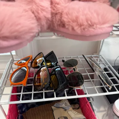 Sale Photo Thumbnail #103: Selection of modern fashion sunglasses displayed on a white wire organizer, with plush pink slippers resting on the shelf above.