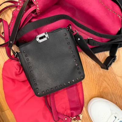 Sale Photo Thumbnail #57: Collection of handbags including a black studded crossbody bag resting on top of a bright pink studded tote, a LeSportsac strap, and a pair of white sneakers on a hardwood floor.