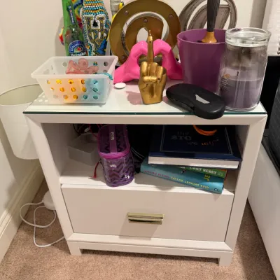Sale Photo Thumbnail #80: White single-drawer nightstand with brass hardware, featuring open shelf storage and a glass-top surface holding various decor, books, and home accessories.
