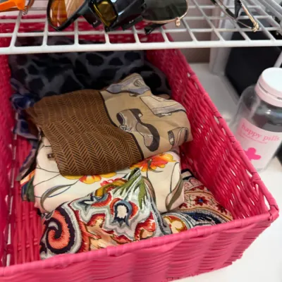 Sale Photo Thumbnail #104: Collection of assorted vintage and contemporary fashion scarves neatly folded inside a bright pink woven storage basket, with various sunglasses resting on a wire shelf above.